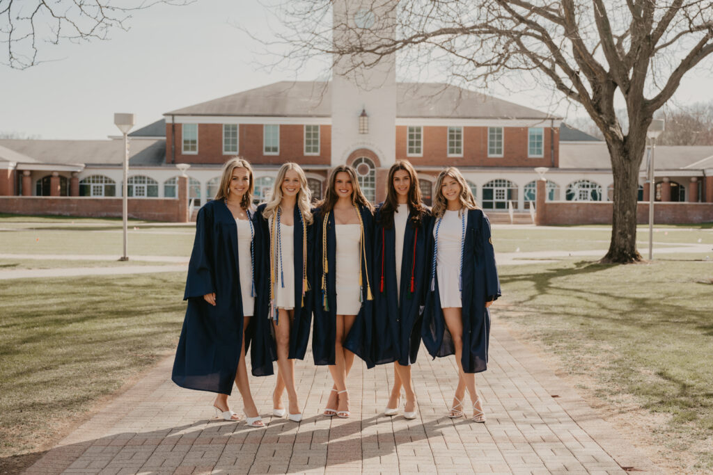 A group of five friends and Quinnipiac graduates wearing white dresses and navy gowns, posing together in front of iconic campus architecture to document their shared journey.