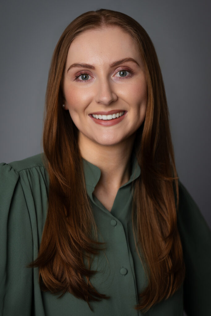 A polished and confident professional headshot of Caroline smiling in a green blouse, photographed with clean studio lighting and a timeless gray backdrop.