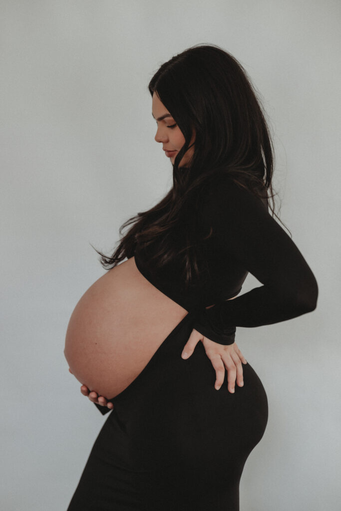 A black and white inspired profile shot of a pregnant woman in a black dress, highlighting the "beauty of motherhood" with clean, minimalist studio lighting.