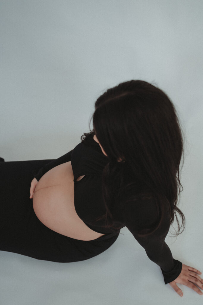 A unique high-angle shot of a pregnant woman sitting on the studio floor in a black dress, highlighting the "beauty of motherhood" through artistic perspective.