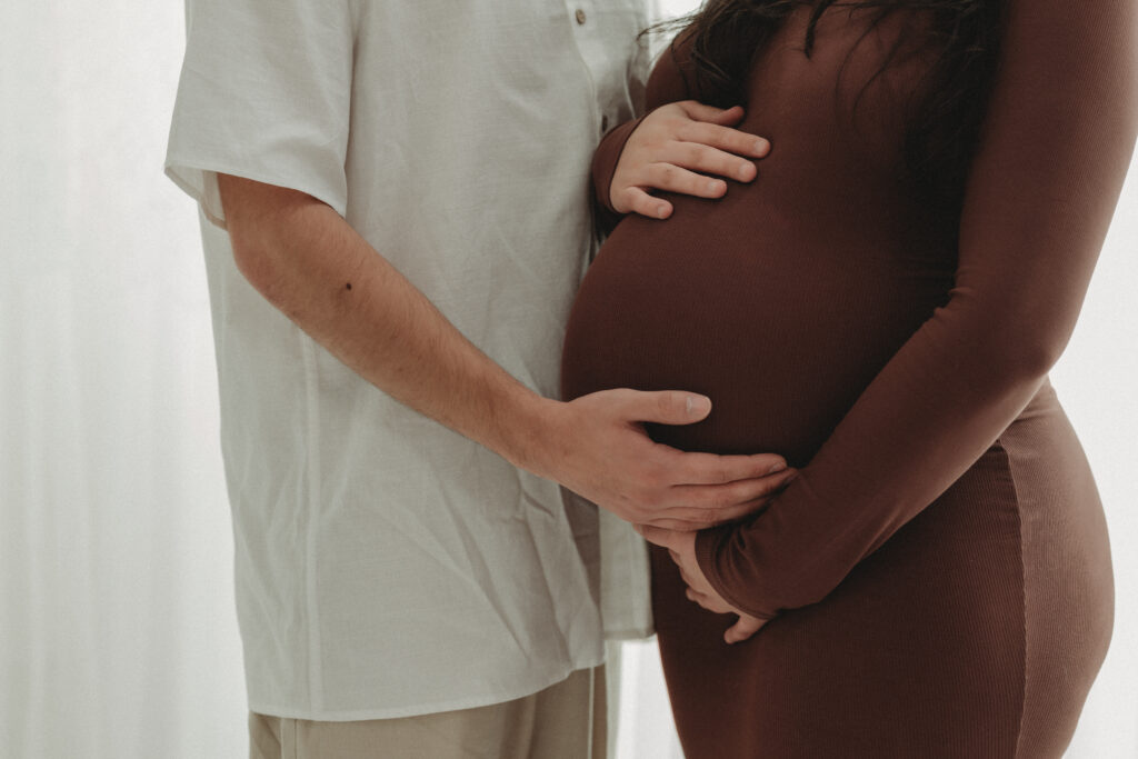 A close-up detail shot of a couple’s hands resting on a maternity baby bump, emphasizing the anticipation and personal connection of the season.