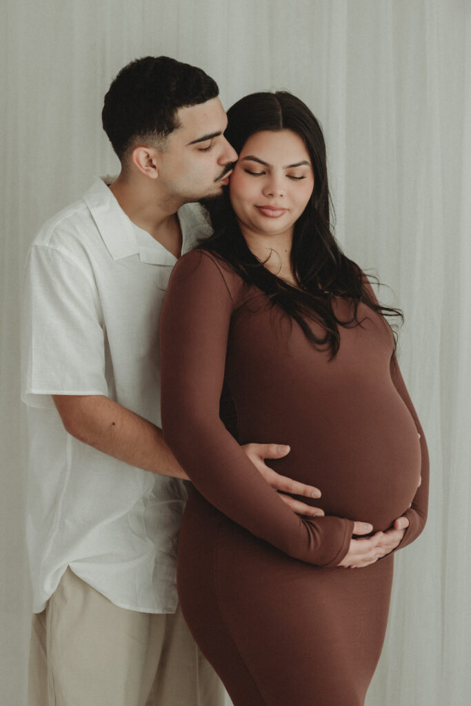 A father-to-be embracing the mother-to-be from behind and kissing her cheek, capturing the emotional connection before welcoming their baby.