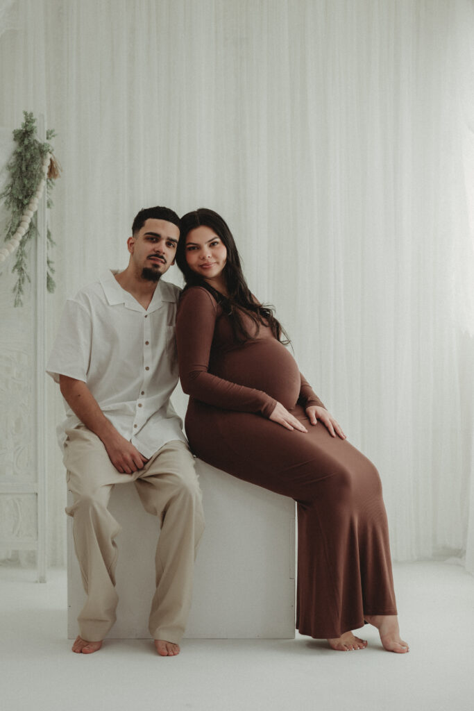 A couple posing together in a bright North Haven studio; the mother-to-be sits on a white block in a long brown gown while the father-to-be sits beside her.