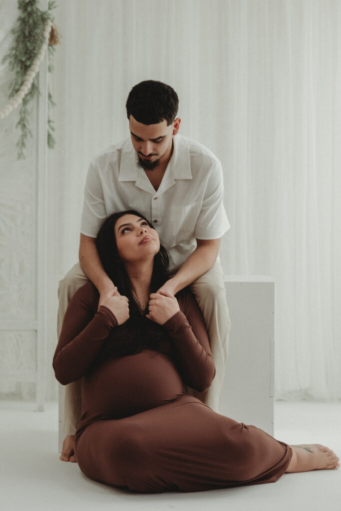 A seated maternity portrait featuring carefully guided posing that creates an elevated editorial feel, with the couple looking at each other against a soft white backdrop.