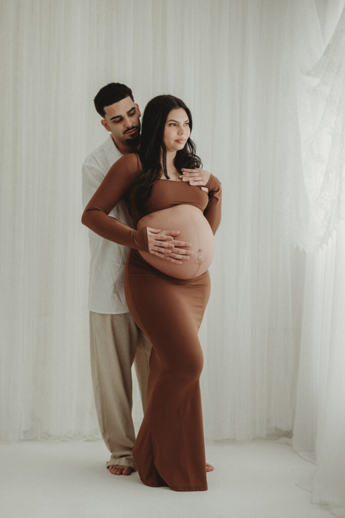 A full-length portrait of a couple standing in a light-filled studio; the mother-to-be wears a flowing brown dress while the father-to-be embraces her from behind.
