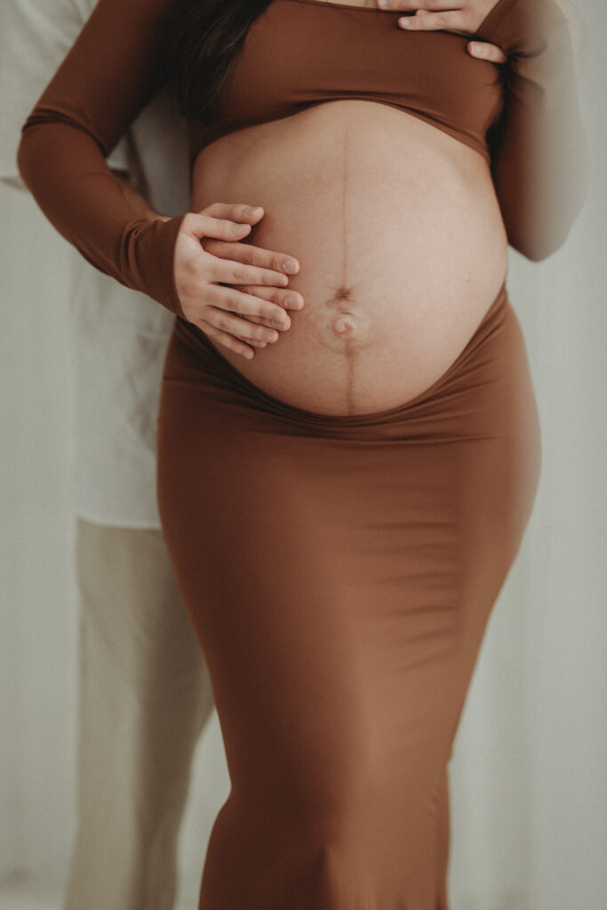 A close-up shot focusing on the maternity bump and the couple's hands, showcasing the "softness and strength" of this season in a lifestyle studio setting.