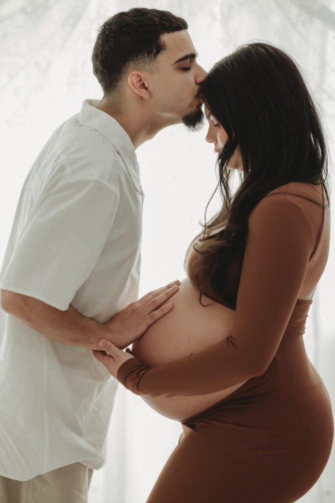 A tender moment of a father-to-be kissing the mother-to-be's forehead while they both place hands on her baby bump during a studio session in North Haven, CT.