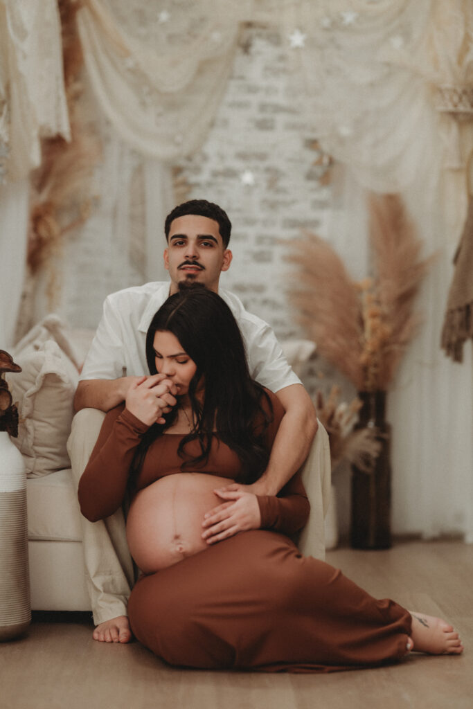 A romantic maternity portrait of a father-to-be kissing the hand of the mother-to-be, who is wearing a brown form-fitting dress in a light-filled lifestyle studio.