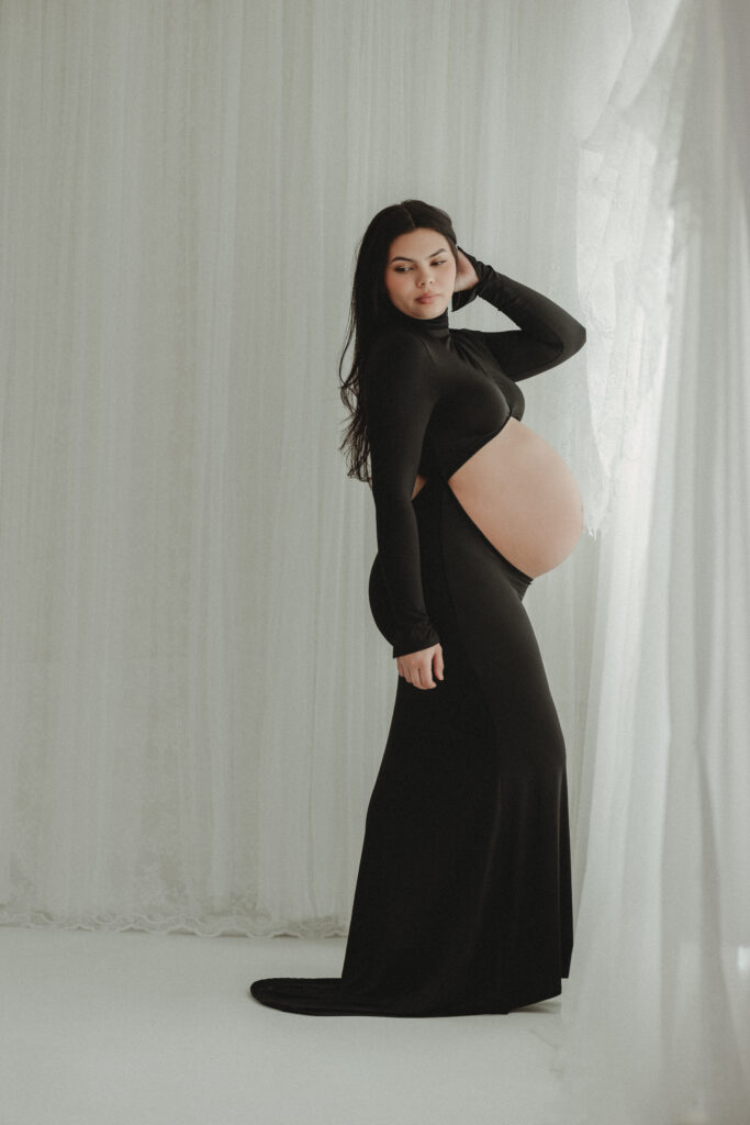 A striking editorial-style portrait of a pregnant woman posing in a long black dress against soft white curtains, capturing a timeless and emotional silhouette.