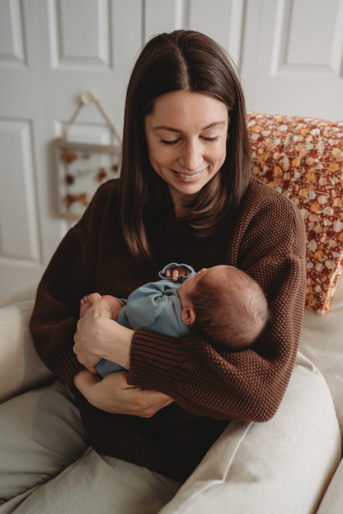A mother in a brown sweater sitting in a nursery chair and smiling down at her newborn baby, reflecting a calm and authentic lifestyle moment.
