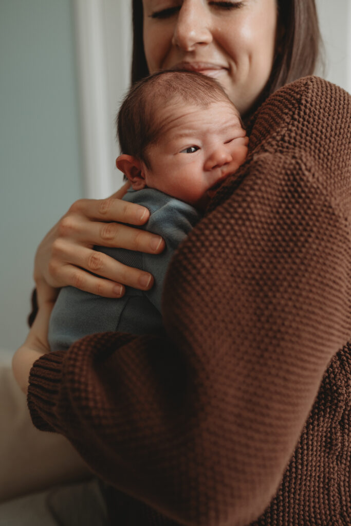A mother wearing a cozy brown sweater cradling her newborn baby against her shoulder, reflecting a calm and heartfelt lifestyle moment at home.