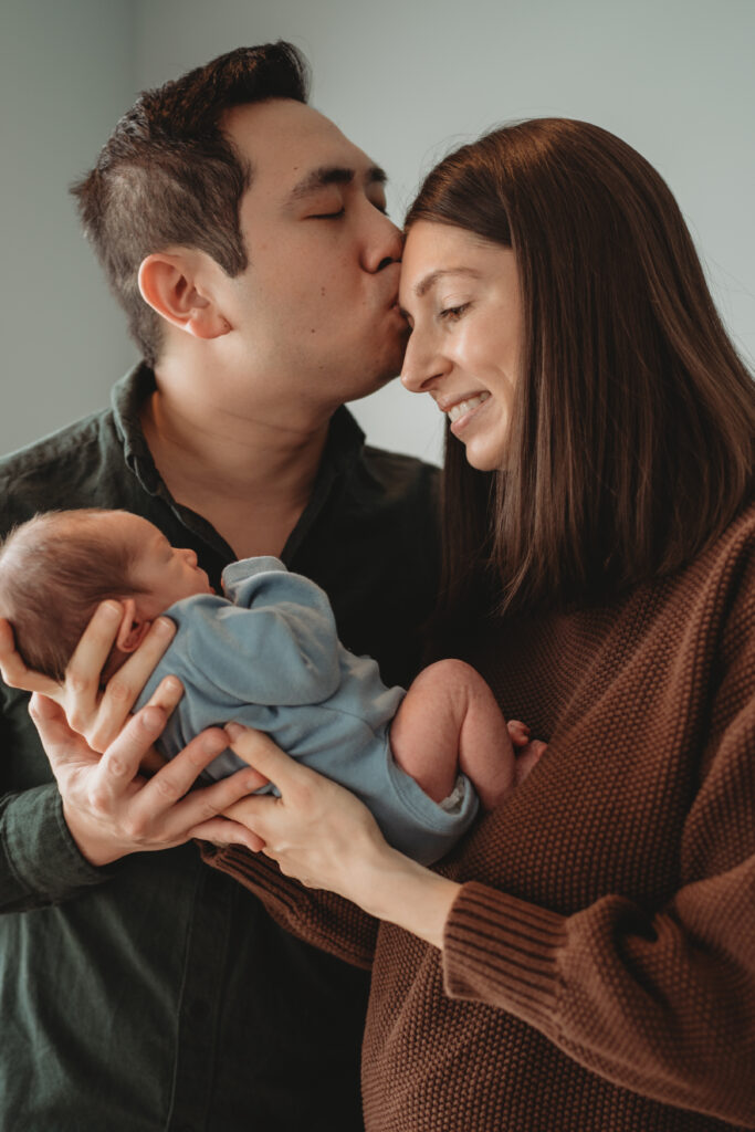 A father kissing his wife's forehead while they both hold their newborn baby, capturing a beautifully authentic moment in their light-filled North Haven home.