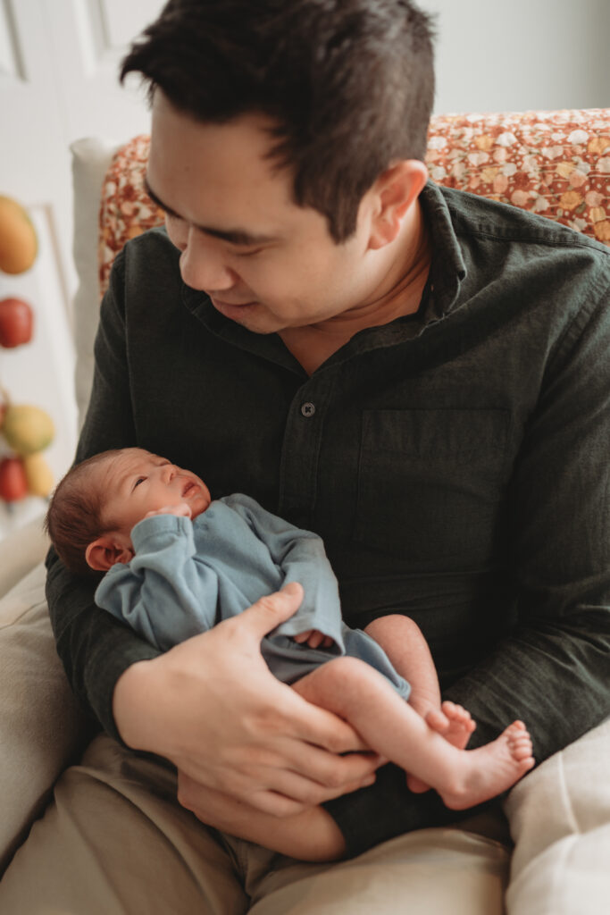 A father sitting in a nursery chair and looking down lovingly at his newborn baby, who is dressed in a soft blue outfit.