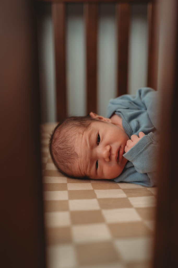 A newborn baby lying on a checkered crib sheet, viewed through the wooden slats of the crib, showcasing the thoughtfully prepared nursery space.