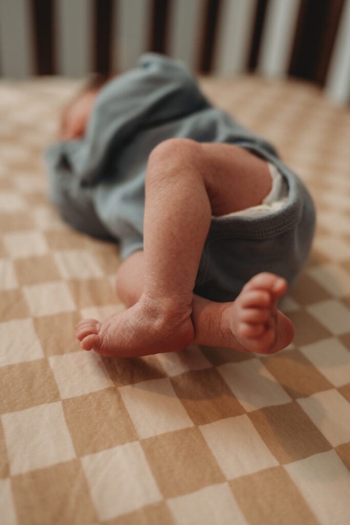 A close-up of a newborn's tiny legs and feet as they lie on a tan checkered crib sheet, documenting the precious details of this new season.