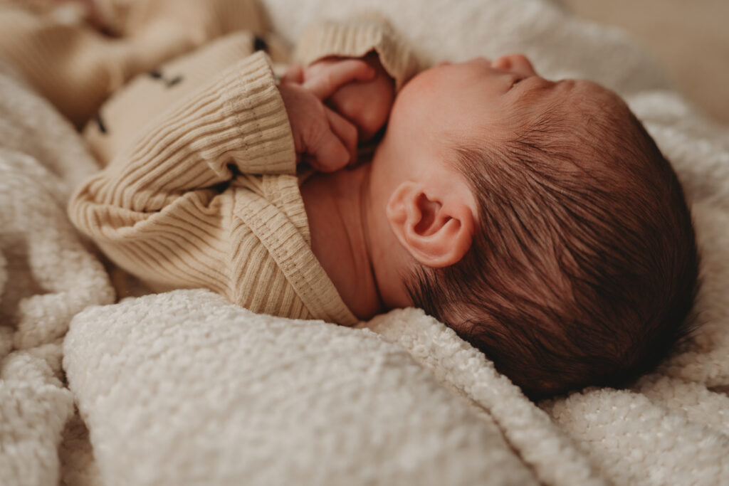 A macro detail shot focusing on a newborn baby's ear and soft hair while resting on a textured cream blanket during an in-home session.