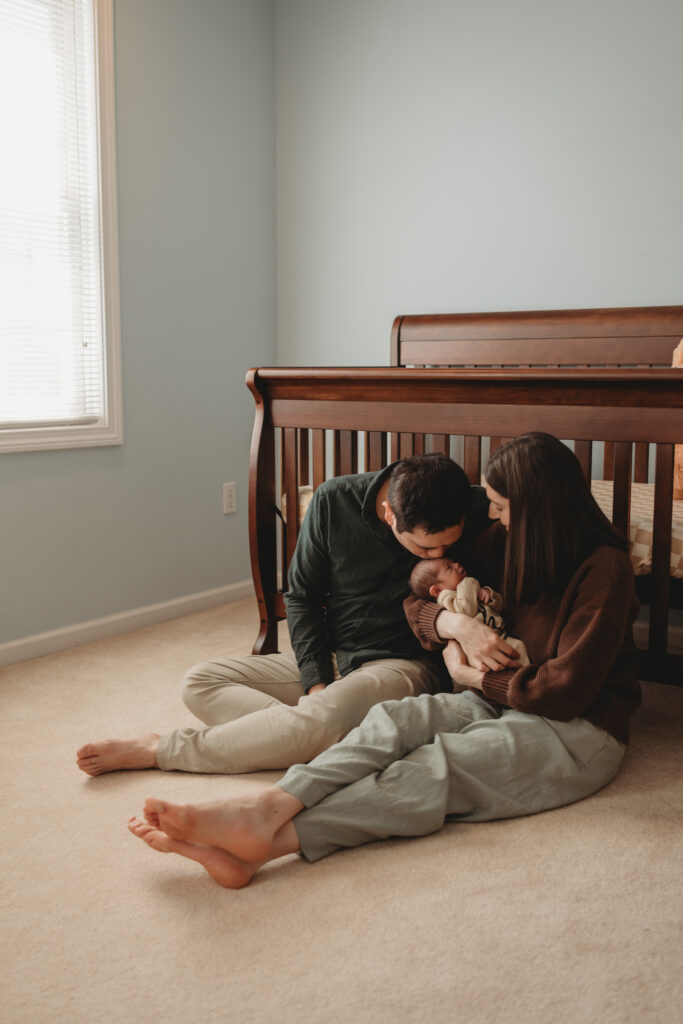 A father and mother sitting on their nursery floor in North Haven, CT, gently cuddling their newborn baby during a cozy lifestyle session.