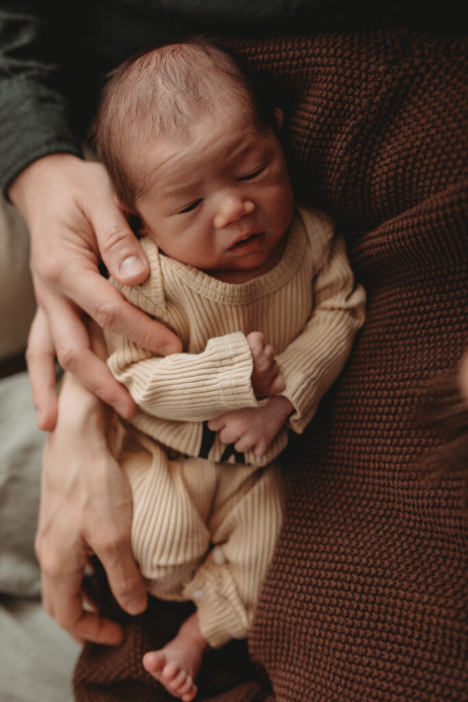 A close-up portrait of a newborn baby wearing a tan ribbed outfit, held securely in their parent's hands to capture "tiny details" and connection.