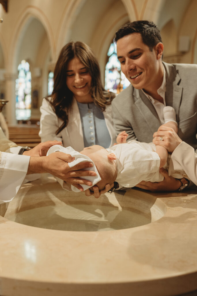 A meaningful moment of the priest performing the blessing at the font while the parents look on with smiles.