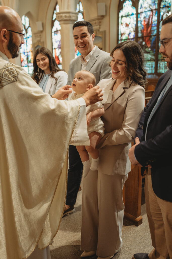 The priest anointing the baby while the mother holds him and family members watch, capturing a key tradition of the baptism celebration.