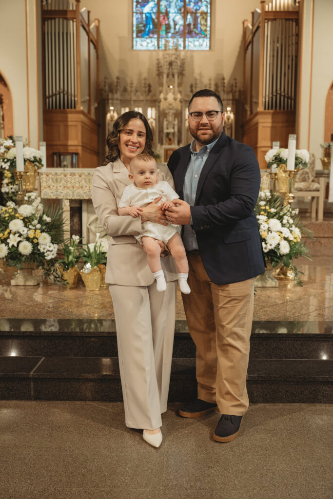 Proud parents standing at the church altar holding their baby after the baptism, surrounded by elegant floral arrangements and stunning architecture.