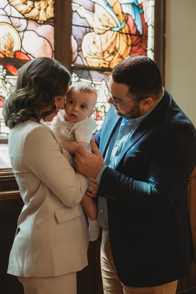 A father and mother looking lovingly at their baby while standing in front of colorful stained glass windows that provide warm, natural light.
