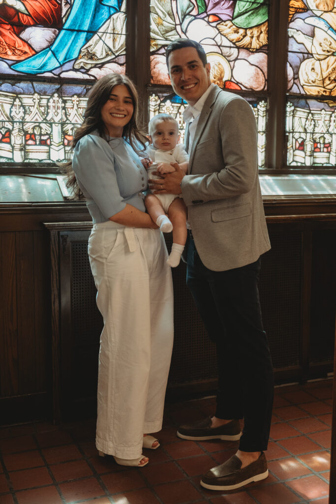 A smiling godfather and godmother holding the baby in front of a stained glass window, documenting the special bond and connection of the day.