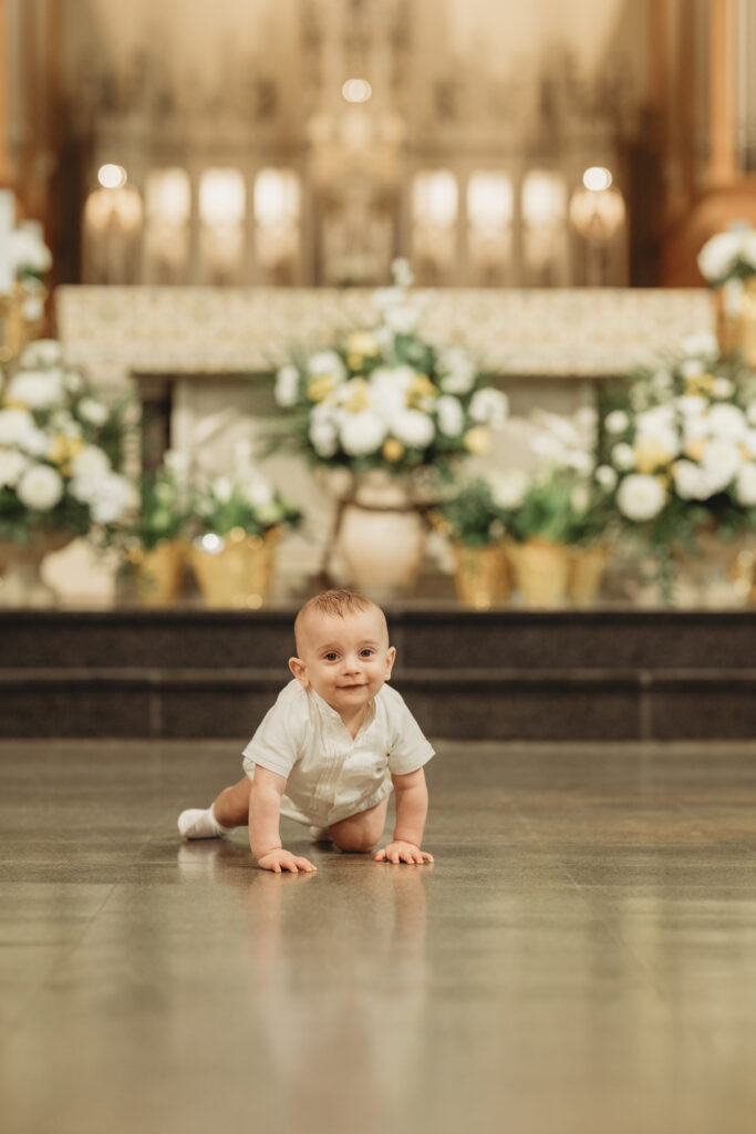 A joyful baby boy in his white baptismal outfit crawling toward the camera on the church floor, with a blurred view of the floral-decorated altar in the background.