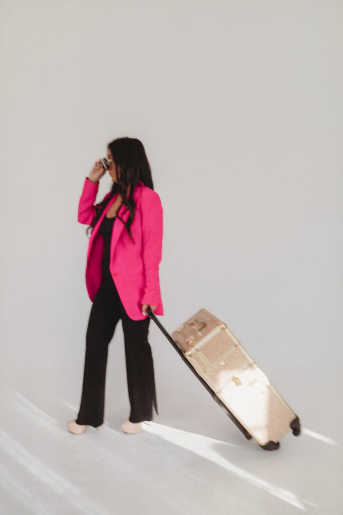 Sarah Khan in a bold pink blazer pulling a gold suitcase, representing her mobile spray tan and beauty service during a branding session at The Studio at 100.