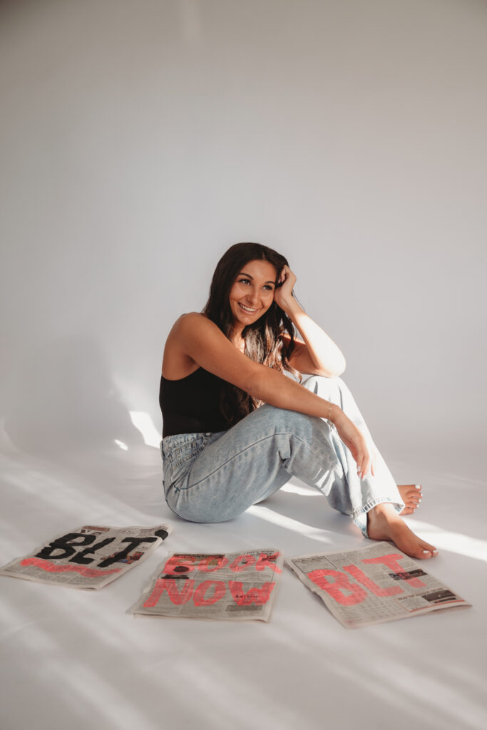 Sarah Khan sitting on the floor in a black tank top and jeans, surrounded by custom BLT newspapers, capturing a "modern and elevated" visual identity.