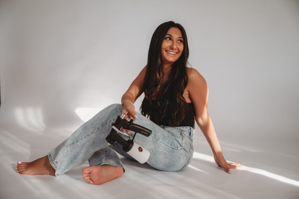 A smiling Sarah Khan holding her professional spray tan machine, designed to help her "connect with future clients" through authentic imagery.