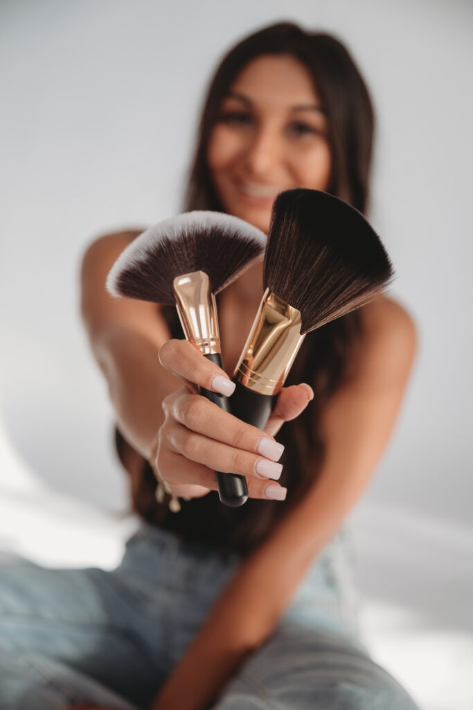 Sarah Khan holding out professional makeup brushes toward the camera, emphasizing the "empowering beauty energy" of her brand.