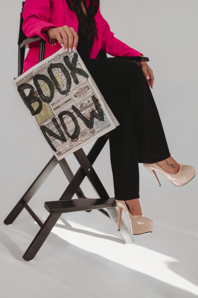 Sarah Khan sitting in a director's chair holding a custom newspaper that says "BOOK NOW," creating elevated imagery for her business launch.