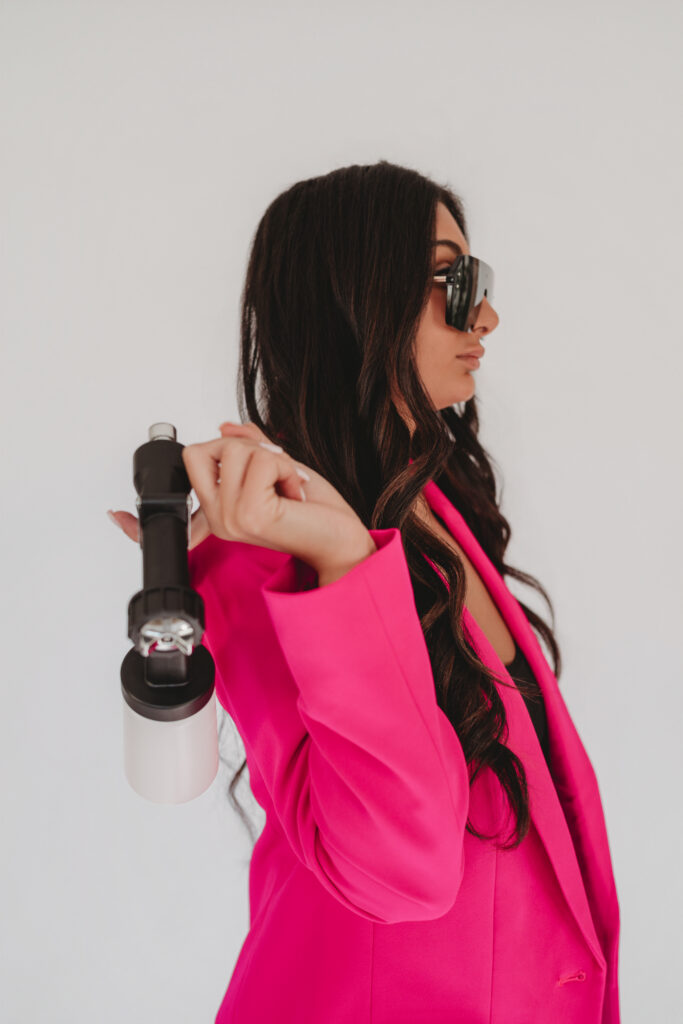 A profile shot of Sarah Khan in a pink blazer holding her professional mobile spray tan equipment, intentionally designed for her online visual identity.