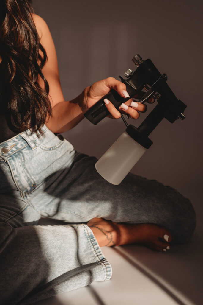 A detailed shot of Sarah Khan holding her spray tan equipment while sitting in a light-filled studio, highlighting the tools of her trade.