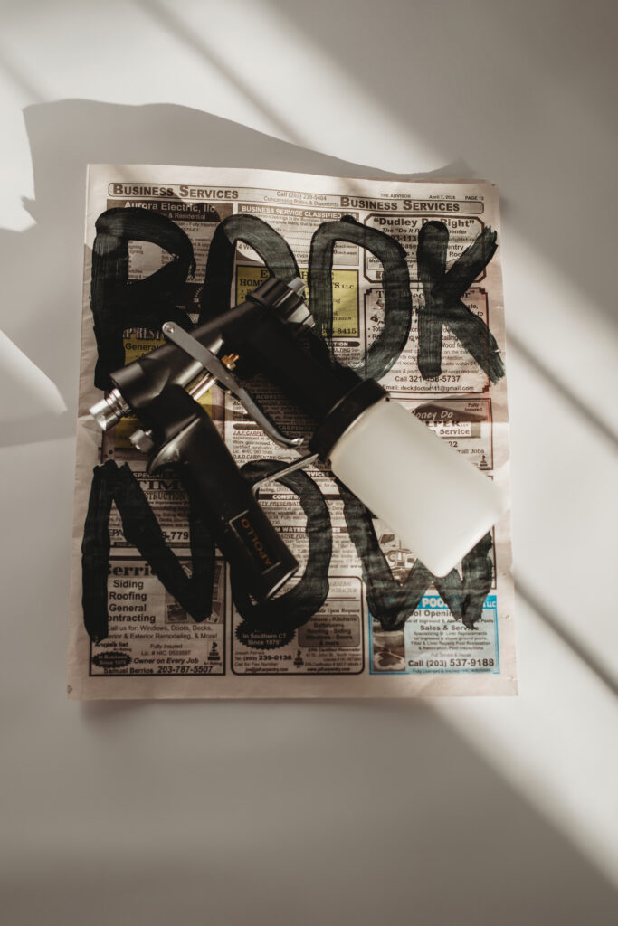 A high-angle shot of a spray tan machine resting on a custom newspaper that says "BOOK NOW," creating "intentionally designed content" for her business launch.