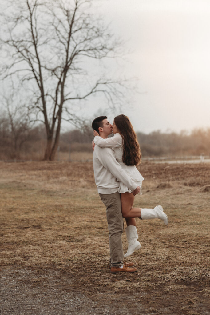 Joe lifting Savannah for a romantic kiss in an open field near the shoreline, featuring timeless sunset tones and a relaxed atmosphere