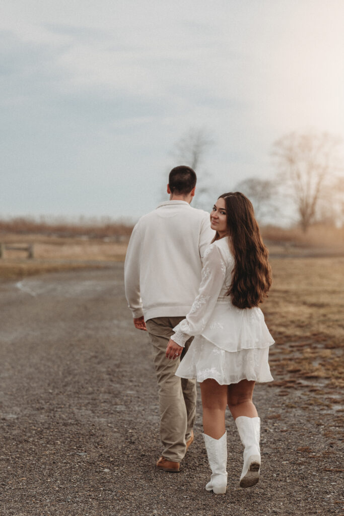 Savannah looking back over her shoulder while walking with Joe along a path, showcasing her white dress and boots against the coastal landscape.