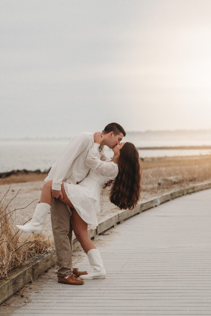 A romantic moment of Savannah and Joe kissing on the boardwalk at Silver Sands State Park in Milford, CT, with the ocean in the background.