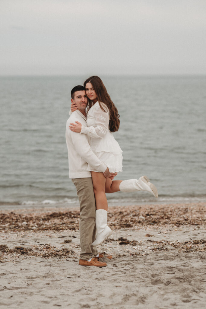 Joe lifting Savannah while they pose on the sandy beach at Silver Sands, showcasing the coastal beauty of this popular Connecticut engagement location.