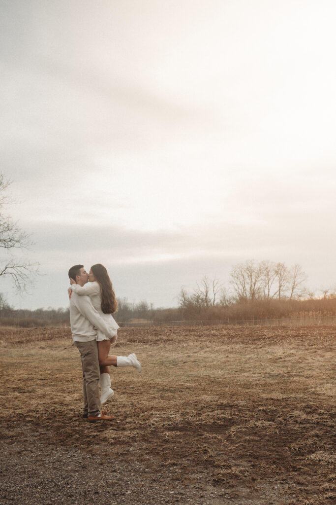 Joe lifting Savannah for a kiss in an open field, highlighted by the timeless sunset tones and soft natural light of Milford, CT.