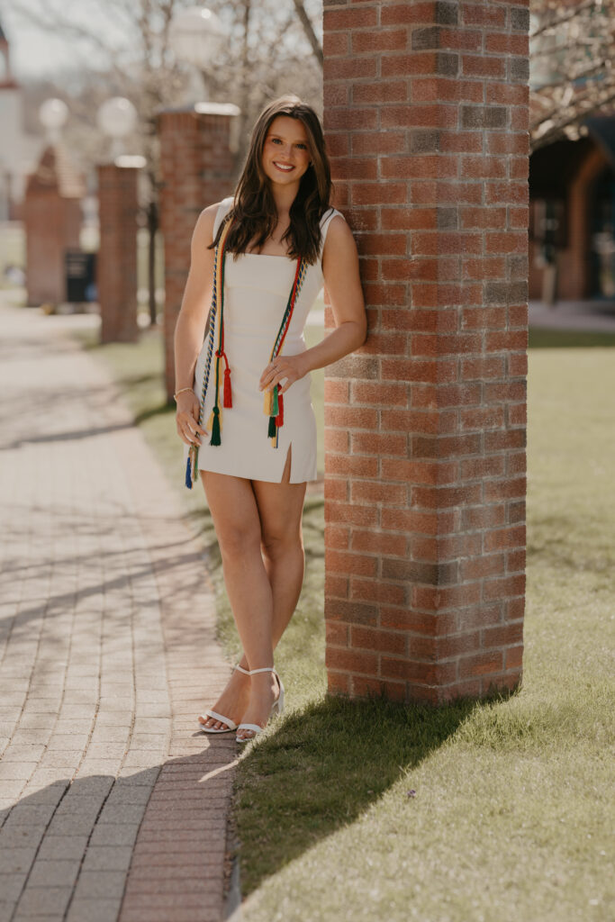 A smiling Quinnipiac graduate leaning against a brick pillar on campus, wearing colorful graduation cords that highlight her academic achievements.