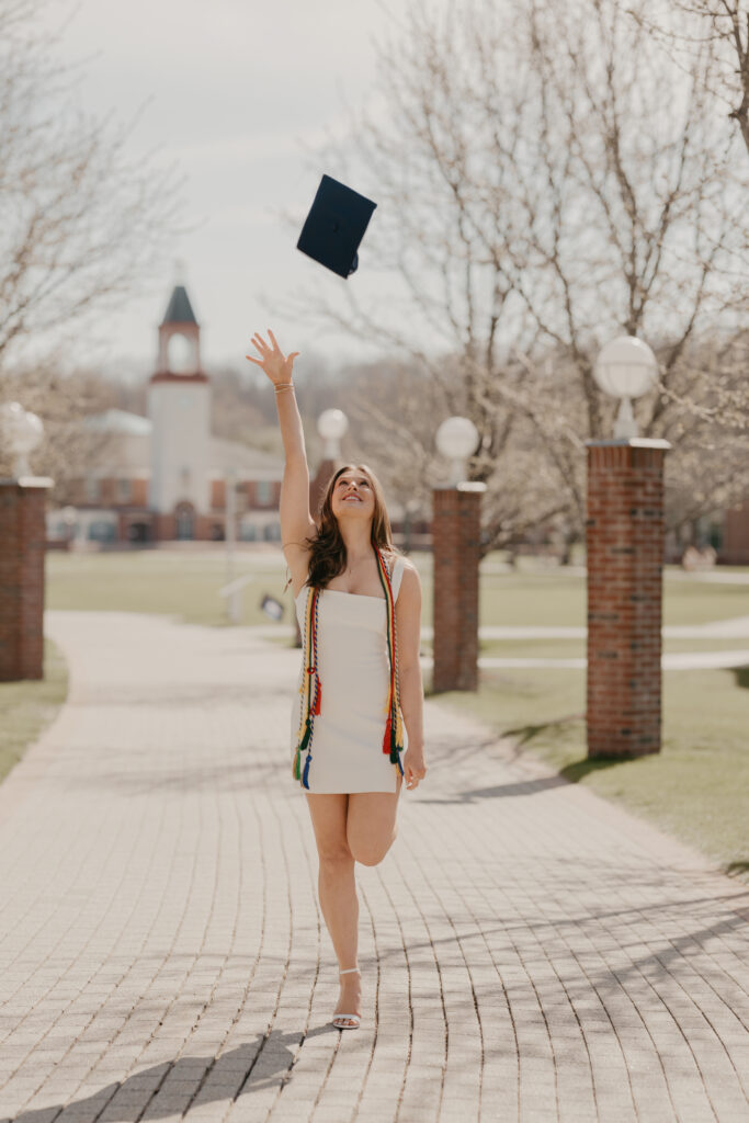 A joyful Quinnipiac graduate throwing her cap into the air on a campus walkway, capturing the excitement and genuine connection of graduation morning.