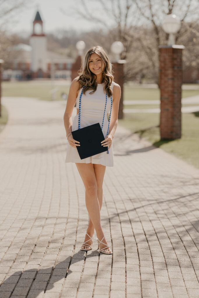 A Quinnipiac University graduate in a white dress and graduation cords standing on a brick campus path, holding her navy graduation cap in the soft early morning light.