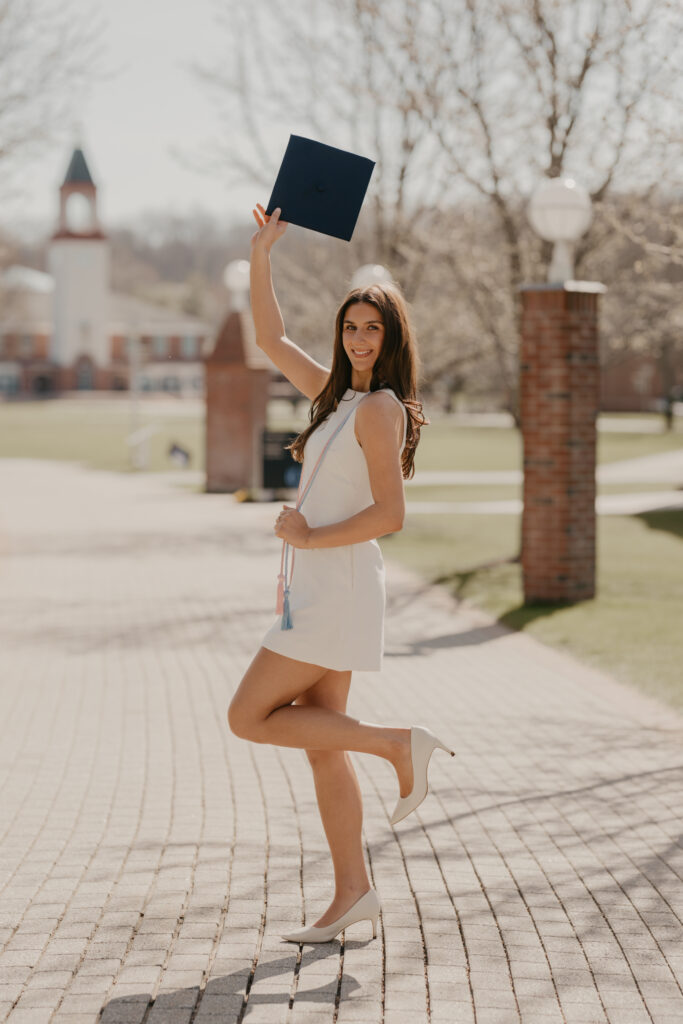 A graduate posing playfully with one foot up and her graduation cap held high, celebrating her milestone on the Quinnipiac campus in Hamden, CT.