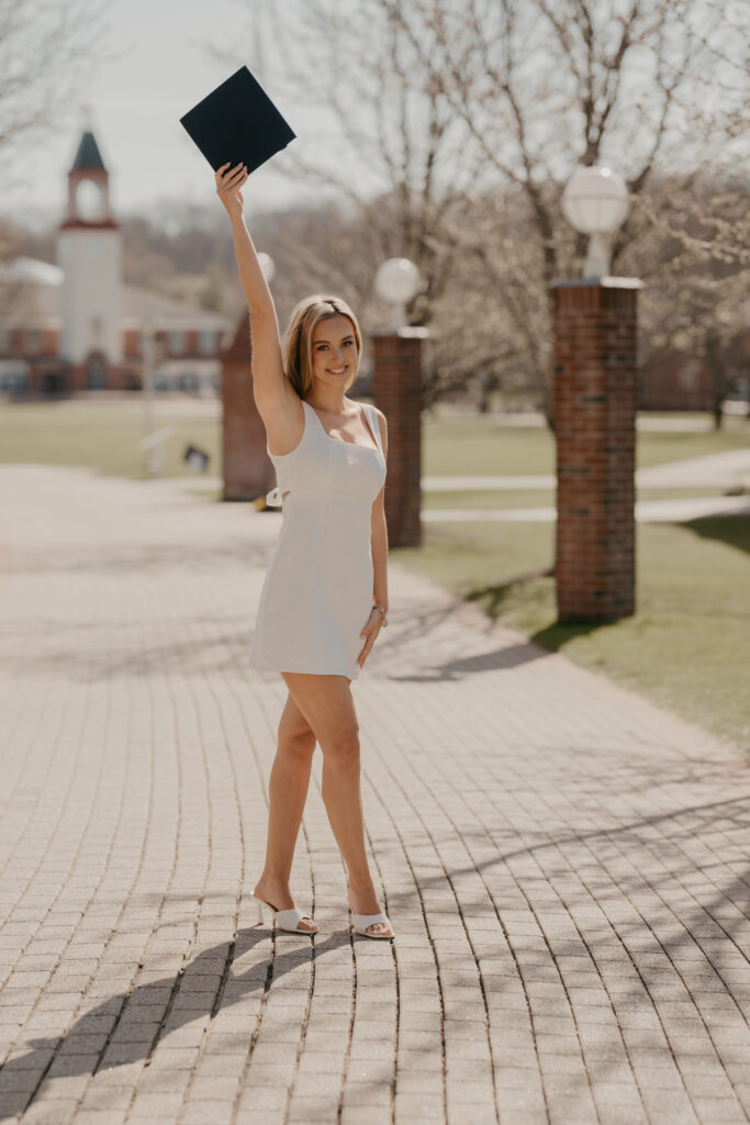 A graduate in a white dress standing on a campus path and raising her graduation cap high in the air, celebrating her graduation chapter.