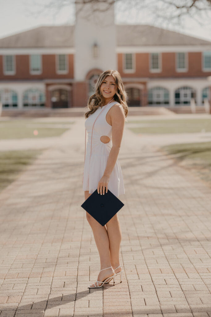 A Quinnipiac graduate posing with her cap on a quiet campus path, highlighted by the glowing early morning light and beautiful school architecture.