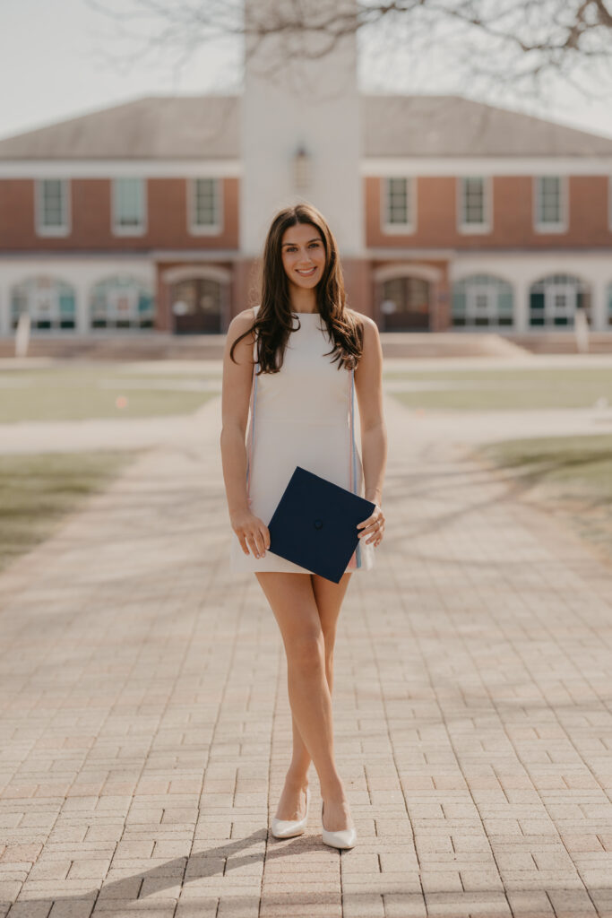 A graduate in a sleeveless white dress holding her navy graduation cap, creating a timeless and professional milestone portrait at Quinnipiac University.