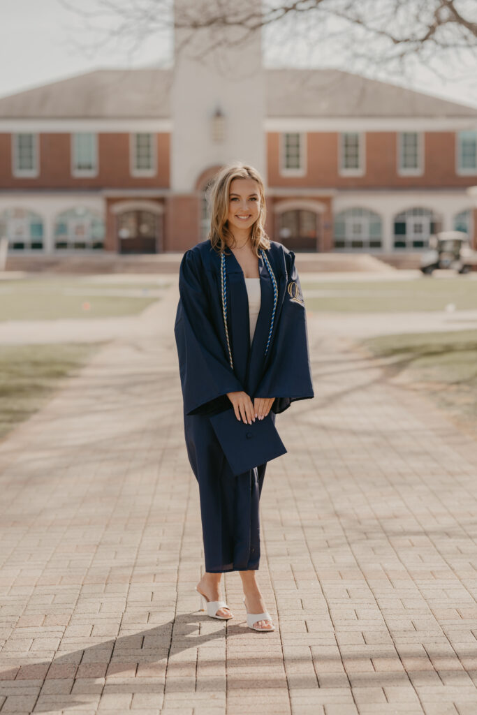 A Quinnipiac University graduate posing in her navy graduation gown and white heels on a sun-drenched brick walkway in Hamden, CT.