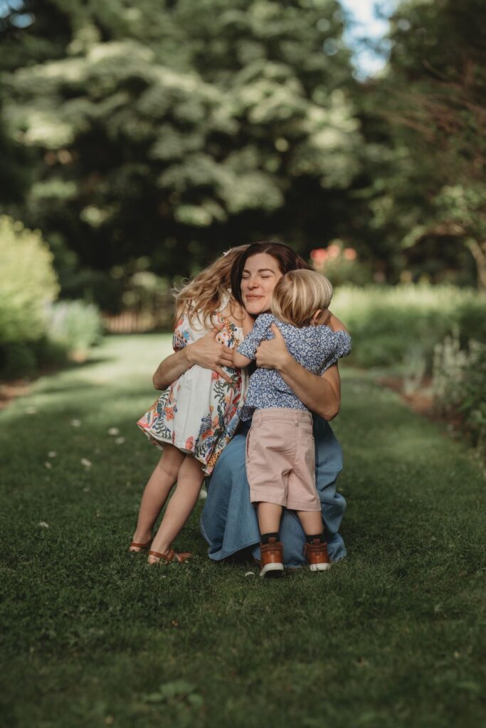 A mother smiling warmly as she embraces her young daughter and toddler son during a golden hour family session in a lush Connecticut garden